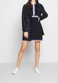 Trench-coat bleu marine porté sur une robe noire avec un col rayé multicolore, assortie à des baskets blanches. Tissu lisse avec des boutons en accent.