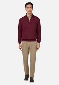 Burgunderfarbener Zip-Pullover, beigen Hosen und dunklen Sneakers. Der Pullover hat gerippte Bündchen und Saum, eine glatte Textur; die Hose hat eine taillierte Passform.