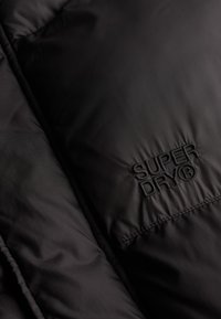 Zwart gewatteerde stof met "SUPERDRY®" geborduurd in zwarte draad aan de rechterkant.