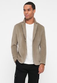 Beige kangaskoots, millel on kaelus, kaks eesmist taskut ja ühe nööbi sulgemine. Tekstureeritud kangas, kohandatud sobivus, kombineeritud valge särkiga.