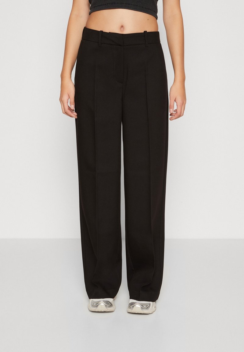 Personne portant un pantalon noir taille haute à jambes larges, associé à des baskets blanches et un top court noir, debout devant un fond blanc.