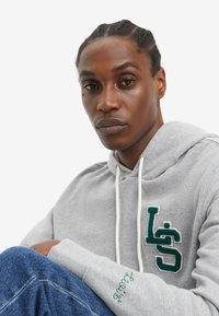 Άνδρας με πλεξούδες που φοράει ανοιχτό γκρι φούτερ με σκούρο πράσινο λογότυπο "LS" και μπλε τζιν, καθισμένος μπροστά σε λευκό φόντο.