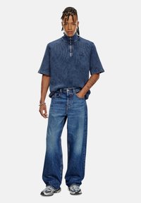 Polo en tricot bleu avec un col zippé, associé à un jean bleu clair ample et des baskets argentées. Coutures simples, sans motifs.