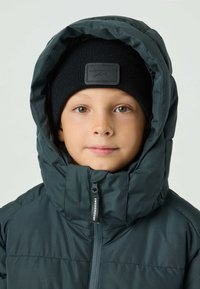 Ocieplana ciemnozielona kurtka z wysokim kołnierzem i zamkiem błyskawicznym, w połączeniu z czarną dzianinową czapką z fakturowaną łatą.