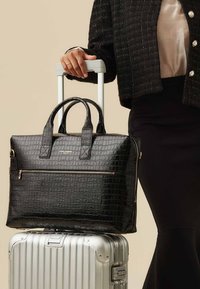 Sac en cuir noir embossé crocodile avec double poignées, poche avant zippée et quincaillerie dorée, reposant sur une valise argentée.