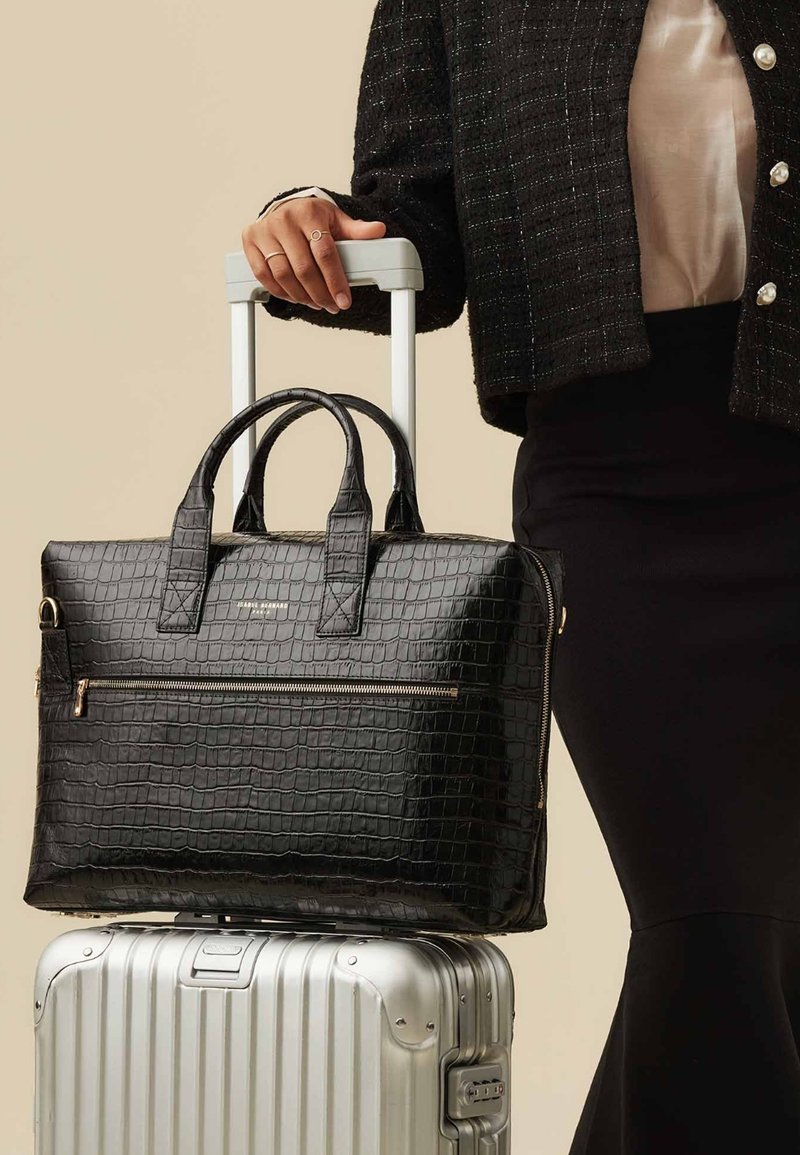 Sac en cuir noir embossé crocodile avec double poignées, poche avant zippée et quincaillerie dorée, reposant sur une valise argentée.