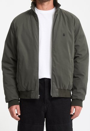 Homme portant une veste bomber vert olive zippée sur une chemise blanche et un pantalon noir, présenté du cou aux hanches sur un fond uni.