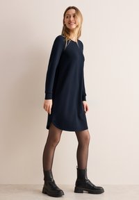 Granatowa sukienka z długim rękawem w luźnym kroju, z zaokrąglonym dołem oraz miękkiego materiału, w połączeniu z czarnymi botkami na kostkę i przezroczystymi czarnymi rajstopami.