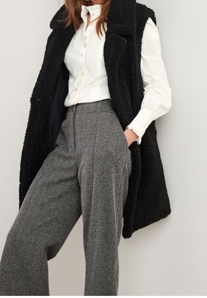 Gilet sans manches noir et texturé sur une chemise blanche à boutons ; pantalon large à motifs pied-de-poule gris avec plis et poches.
