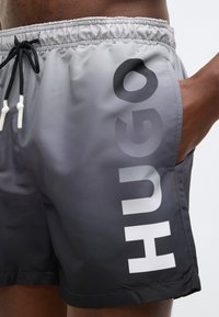 Ανδρικά μαγιό σορτς σε διαβαθμισμένο γκρι, με μαύρη μέση, ρυθμιζόμενο κορδόνι και μεγάλο λευκό λογότυπο "HUGO" στο πλάι.