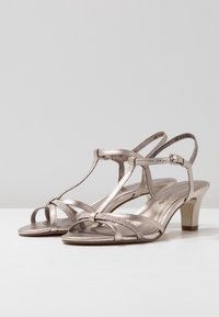 Sandales à talons en métal rose doré pour femme, avec des lanières et fermeture à boucle, à talons blocs de hauteur moyenne sur une surface blanche.