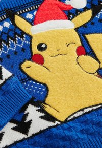 Żółty Pikachu w czapce Świętego Mikołaja, umieszczony na niebieskiej dzianinie z geometrycznym wzorem. Tekstura jest miękka, z prążkowanymi niebieskimi mankietami.