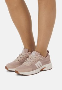 Persona vistiendo zapatillas deportivas de malla ligeras de color rosa claro con cordones blancos y suelas blancas, de pie sobre una superficie blanca.