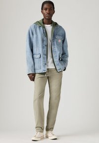 Young man wearing light blue denim jacket over green hoodie, white shirt, beige pants, and white sneakers, standing against plain background.