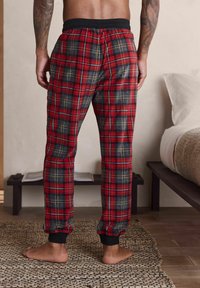 Pantalons de pyjama à carreaux rouges et marine avec une ceinture élastique noire et des bretelles aux chevilles, fabriqués en tissu doux avec une finition texturée.