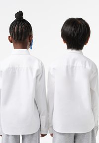 Due bambini indossano camicie bianche con bottoni e maniche lunghe, visti da dietro. Un'acconciatura a treccia e un orecchino blu sono visibili su un bambino.