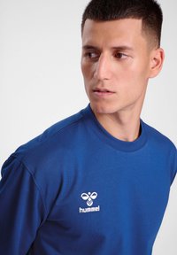 Blå långärmad t-shirt i slät tyg, med rund halsringning och vitt broderat "hummel"-logotyp på vänster bröst.