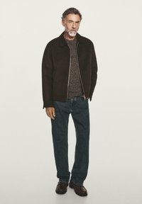 Giacca marrone con zip sopra un maglione a trama, jeans in denim blu e scarpe marroni scure. La figura è in piedi su uno sfondo neutro.