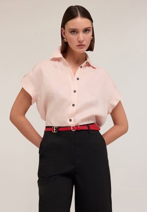 Lichtroze, kortomarm shirt met een kraag en zwarte knopen, gecombineerd met hoge zwarte broeken en een rode riem met gouden beslag.