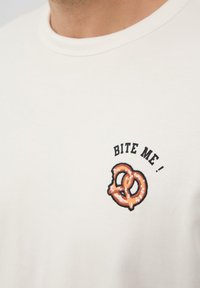 Weißes Baumwoll-T-Shirt mit einem kreisförmigen Brezel-Design und dem Text "BITE ME!" in schwarzer und orangefarbener Stickerei auf der Brust.