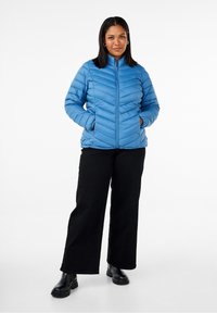 Giacca puffer blu con motivo a chevron, zip frontale, colletto alto e tasche laterali. Abbinata a pantaloni neri a gamba larga e stivali neri.