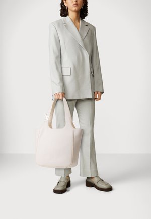 Žena v světle šedém obleku a s platformními mokasínami, držící velkou světle růžovou kabelku na jednobarevném bílém pozadí.