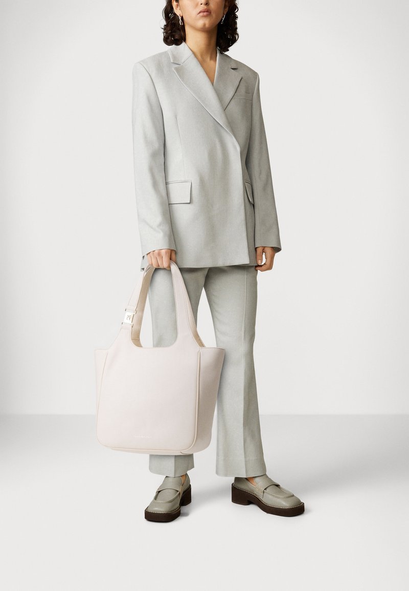 Femme vêtue d'un costume gris clair et de mocassins à plateforme, tenant un grand sac à main rose pâle sur un fond blanc uni.