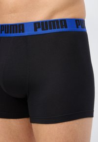 Άνδρας που φοράει μαύρα βαμβακερά μποξεράκια με μπλε ζώνη μέσης όπου επαναλαμβάνονται μαύρα λογότυπα "PUMA", φαίνεται από τη μέση έως το μέσο του μηρού σε πλαϊνή γωνία.