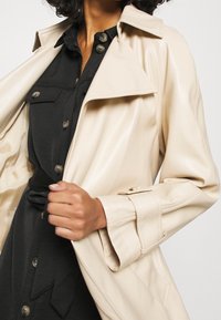 Beige faux leather trench coat with large collar, button details, and wide sleeves. Worn over a black button-up dress.