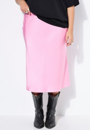 Kobieta w jaskrawo różowej spódnicy midi, czarnym topie, pierścionkach i czarnych kowbojskich butach z ćwiekami, stoi na białym tle.