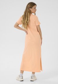 Różowa sukienka maxi z t-shirta w miękkim materiale, z krótkimi rękawami i bocznym rozcięciem, noszona z białymi sneakersami i szarymi skarpetkami.