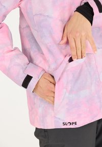 Wasserdichte Jacke mit pinkem und lila abstrakten Muster, ausgestattet mit einer Reißverschlusstasche, verstellbaren Manschetten und einem Logo am Saum, das eine Hand zeigt, die hinein greift.
