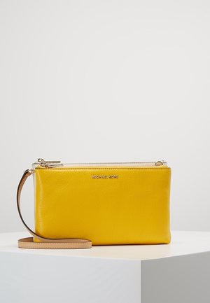 Żółta skórzana bransoletka z beżowym paskiem i złotym zamkiem błyskawicznym, z logo Michael Kors, prezentowana na białym cokole na jednolitym tle.