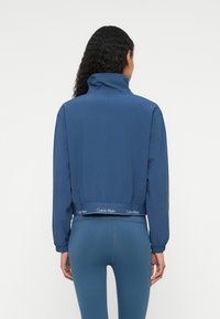 Modrá skrátená bunda s vysokým golierom, elastickými manžetami a pásom s logom "Calvin Klein". Hladká textúra a minimalistický dizajn.