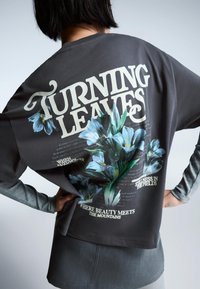 Tricou gri cu maneci lungi, cu un design floral ce include flori albastre și text. Materialul pare moale, cu un croi casual și mâneci suprapuse.