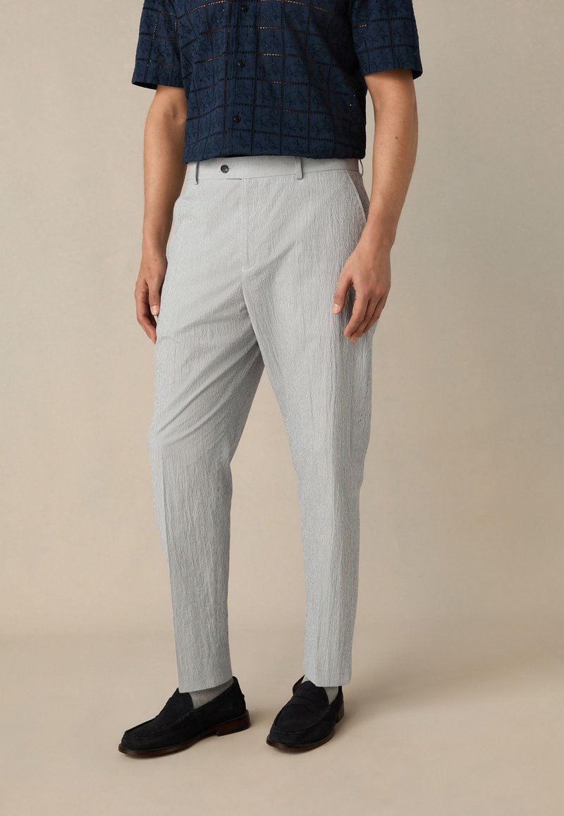 Pantalon gris clair texturé, coupe slim, avec un devant plat et de fines rayures verticales, associé à une chemise foncée à motifs et des mocassins noirs.