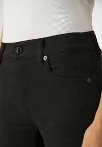Pantalones vaqueros de mezclilla negra con un corte ajustado, que cuentan con un bolsillo para monedas en la parte frontal y un cierre clásico de botón metálico. Textura suave con sutiles acentos de costura.