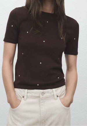 Žena oblečená v tmavě hnědém krátkém tričku s drobnými lesklými nýty, v kombinaci se světle béžovými kalhotami s vysokým pasem, ruce v kapsách.