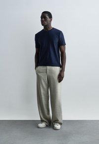 Σκούρου δέρματος άνδρας στέκεται με τα χέρια στις τσέπες, φορώντας μπλέ σκούρο μπλουζάκι, μπεζ παντελόνι με φαρδιά πόδια και λευκά αθλητικά παπούτσια, μπροστά σε απλό φόντο.