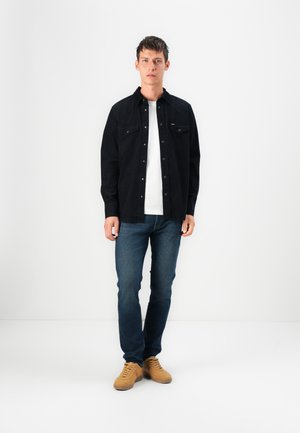 Black long-sleeve shirt with buttoned pockets, worn over a white t-shirt, paired with dark blue jeans and light brown shoes.