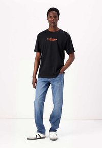 Sort bomulds T-shirt med orange tekst "Creative Works" på brystet, parret med lys blå denim jeans og hvide sneakers med sorte detaljer.