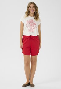 T-shirt crème à manches courtes avec un graphique floral rouge. Associé à un short rouge, celui-ci possède une taille élastique et un cordon de serrage.