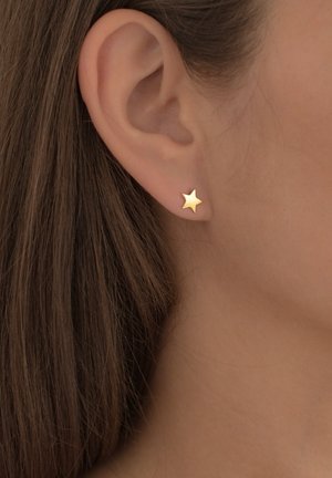 Goldene sterneförmige Ohrringe, die an einem Ohr einer Person getragen werden. Sie haben eine glatte Oberfläche und ein einfaches Design, das das Licht dezent reflektiert.