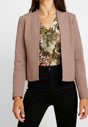 Femme portant un blazer court taupe sur un chemisier à motif vert, marron et rose avec un col en V, et un pantalon noir taille haute.