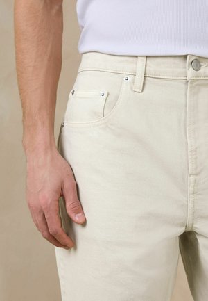 Close-up of a person’s left hand resting beside light beige denim jeans with front pocket and rivet details, wearing a white ribbed shirt.