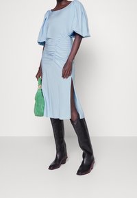 Abito azzurro con maniche a sbuffo e spacchi laterali, realizzato in tessuto morbido. Abbinato a stivali neri sopra il ginocchio e una borsa verde.