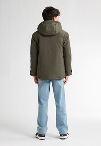 Jachetă căptușită verde măslin cu glugă, cu o textură netedă și manșete elastice, asortată cu blugi drepți de culoare albastru deschis și teniși.