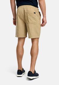 Tan bomuldsshorts med elastisk talje, der har en baglomme og kort kant. Båret med marineblå sneakers og en mørk skjorte.