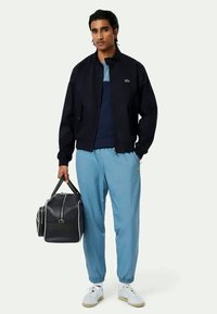 Laivaston pommikaverto, vaaleansiniset housut, valkoiset lenkkarit. Pitelee mustaa duffel-laukkua. Takissa ja housuissa Lacosten logo.