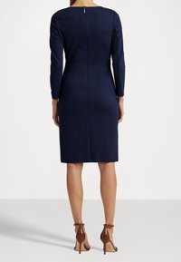 Dopasowana sukienka w kolorze granatowym z długimi rękawami, zamkiem z tyłu i sięgająca do kolan. Noszona ze złamanymi brązowymi sandałami na obcasie.